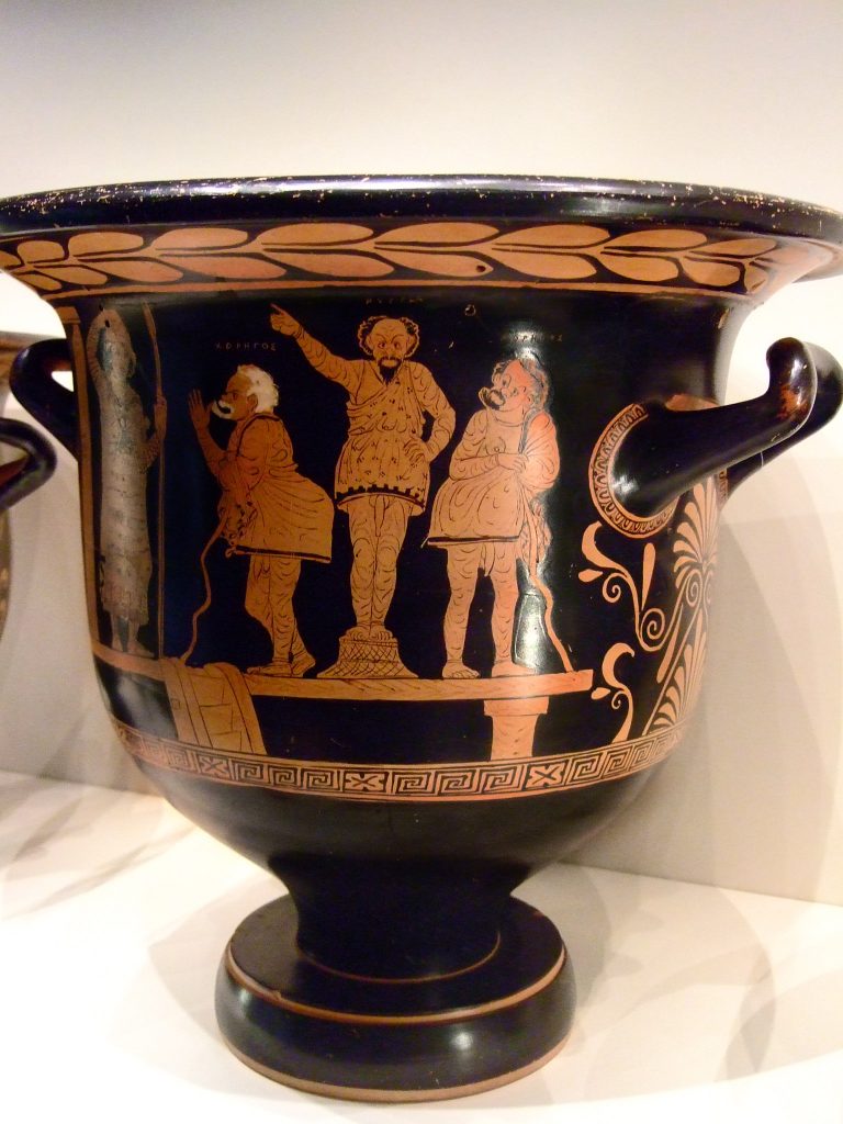 A red-figure painted vase from c. 380 titled, Red-figure Bell Krater with Phlyax Theater scene, depicting two members of the chorus surrounding a protagonist character.