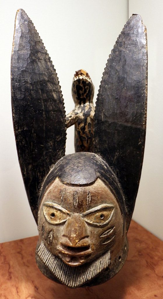 This egungun mask has long ears with a lizzard in between which seems to be controlling the figure below. The figure also has warrior lines carved into the cheek and forehead with wide eyes and plump lips. 