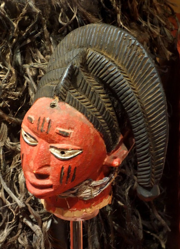 This egungun mask depicts a red figure with what looks like brown warrior stripes across the forhead and cheek. The lips are plump and the eyes are wide. The figure is also wearing a brown headdress that droops down to the right with immaculate line carvings throughout.  