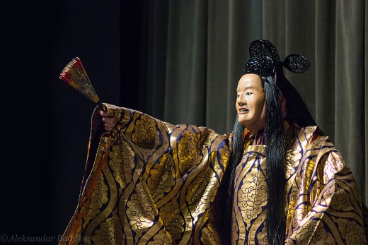 A noh actor turned towards the left of the image. He is holding his right arm up, which is holding a red and yellow fan. He is wearing a blue, tan, and yellow kimono with a repeated cloud-like motif, as well as a long black wig and a black headpiece. He is wearing a mask which has a fairly neutral expression.